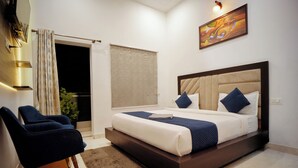 A modern bedroom with a large bed, a seating area with chairs, a window with curtains, and a painting on the wall. - The Nines Hotel & Restro (Haldwani)
