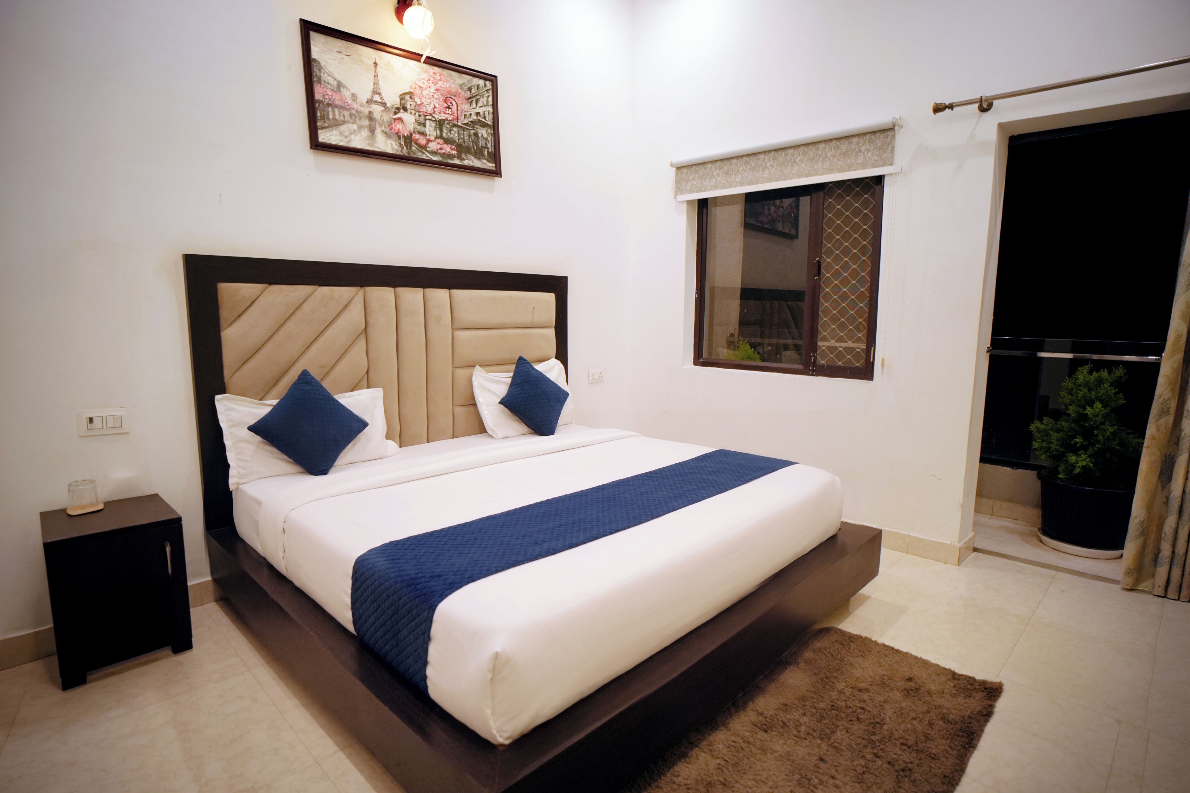A hotel room with a bed, a nightstand, a window with a view, and a painting on the wall.
