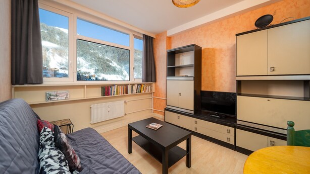 A cozy living room with a sofa, a coffee table, and a TV. There are bookshelves and a kitchen area with cabinets and a stove. - Charming apartment for 6 people, mountain view, ski slope (Allos)