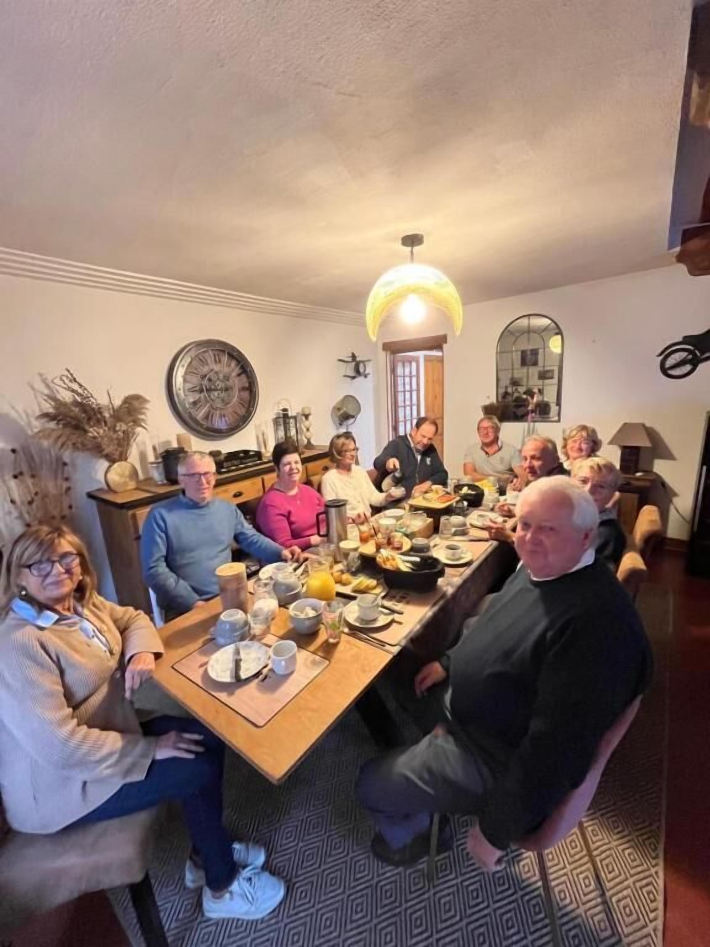 A group of people seated around a wooden dining table in a room with a patterned floor, a large wall clock, and a hanging light fixture.