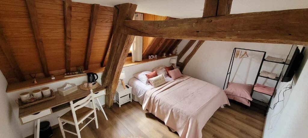 Une chambre confortable située dans les combles, avec un plafond en bois, un lit avec une literie rose, un bureau avec une chaise et une petite étagère où sont posés quelques objets.