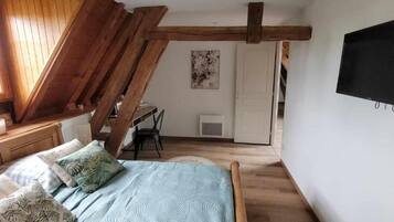 A bedroom with a sloped ceiling, a bed with patterned bedding, a desk with a chair, and a television mounted on the wall.