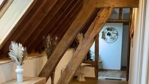 Una escalera de madera conduce a una zona de altillo con un escritorio de madera y un jarrón de flores secas.