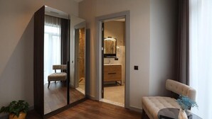 Une chambre d’hôtel avec un plancher en bois, un miroir, un coin salon agrémenté d’un vase de fleurs et une vue sur la salle de bain par une porte ouverte.