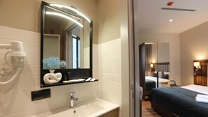 Une chambre d’hôtel moderne avec un grand miroir, un lavabo blanc, une table de chevet et un lit avec une literie aux couleurs sombres.