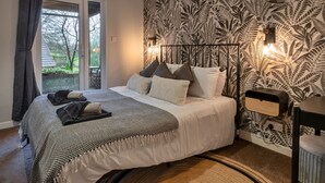 A bedroom with a bed, a window with curtains, a wall with a leaf pattern, and a nightstand with a lamp. - Toblerone Lodge (Callington)