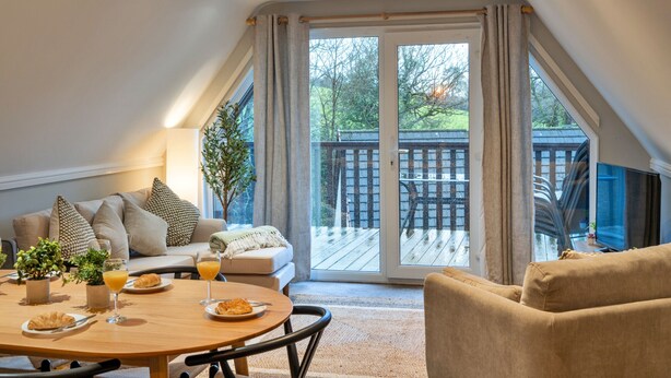 A cozy living room with a dining table set for breakfast, a sofa, and a large window with a view of a wooden deck. - Toblerone Lodge (Callington)