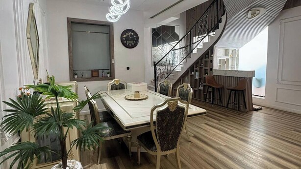 Un espace repas moderne avec un plancher en bois, une table à manger blanche et des chaises. Un escalier avec une rampe noire mène à l’étage supérieur.