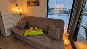 A cozy living room with a sofa, a tray with cups and a teapot, a radiator, a window with a view of boats, and a wall with a framed quote. - ÖKO houseboat Morgenrot with sun terrace & harbor view near Stralsund (Stralsund)