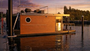 A wooden houseboat with a circular window, a door, and a small deck area. - ÖKO houseboat Morgenrot with sun terrace & harbor view near Stralsund (Stralsund)