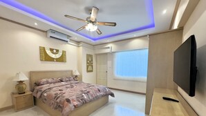 A modern bedroom with a bed, bedside tables, a ceiling fan, an air conditioning unit, a television, and a window with blinds. - Stylish 1-bedroom Corner Condo Near Thepprasit Night Market -keha Condominium (Pattaya)
