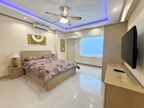 Ein modernes Schlafzimmer mit Bett, Nachttischen, Deckenventilator, Klimaanlage, Fernseher und einem Fenster mit Jalousien.