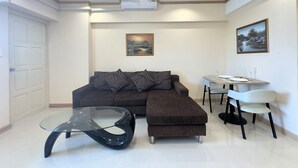 A modern living room with a sectional sofa, a glass coffee table, and a dining area with a small table and chairs. - Stylish 1-bedroom Corner Condo Near Thepprasit Night Market -keha Condominium (Pattaya)