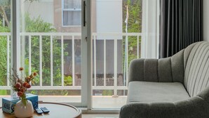 Ein Wohnzimmer mit grauem Sofa, einem runden Holztisch mit einer Vase und Blick auf ein Haus durch eine Glastür.