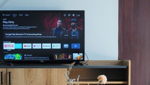 Ein Fernseher zeigt eine Filmempfehlung, eine Fernbedienung, eine Vase mit Blumen und eine Schachtel mit Zeichentrickfiguren auf einem Holztisch.