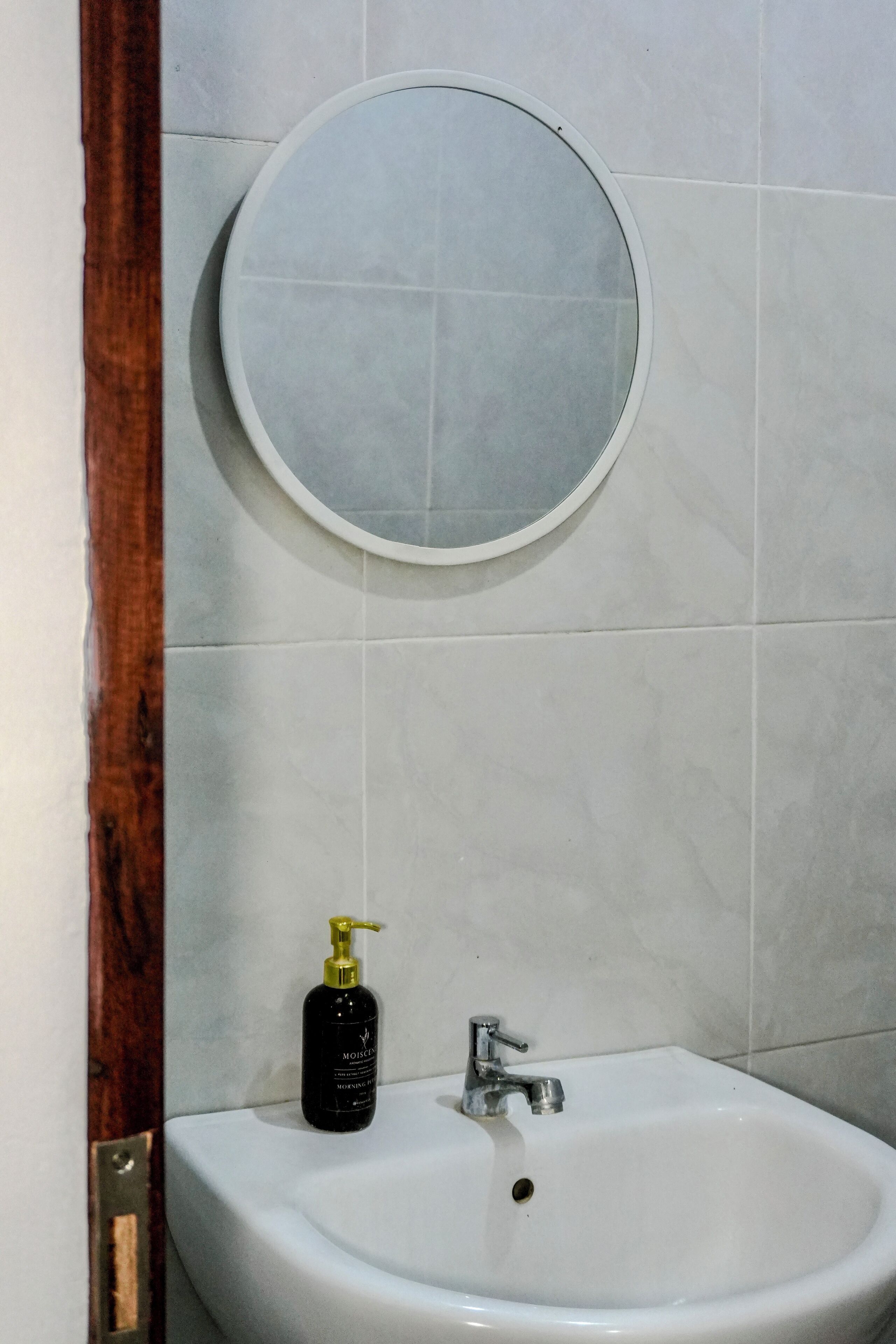 Une salle de bain avec un lavabo blanc, un miroir rond et une bouteille de shampoing.