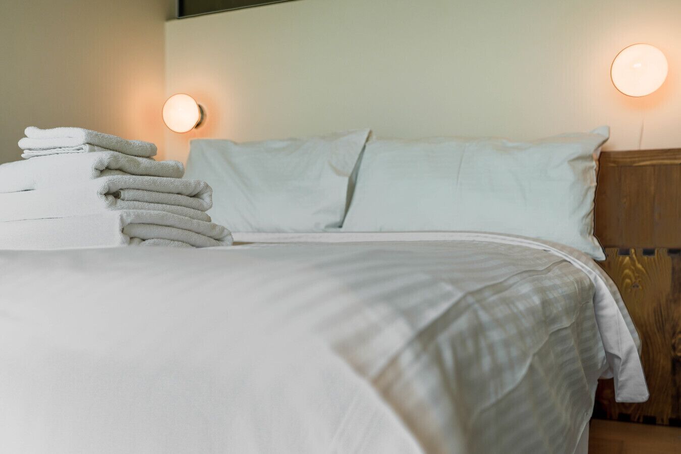 Un lit bien fait, avec du linge de lit blanc, deux lampes fixées au mur et une pile de serviettes pliées.