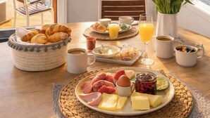 Uma mesa de café da manhã com croissants, ovos, presunto, queijo e frutas, posta sobre uma mesa de madeira com vista para barcos na água através de uma janela.