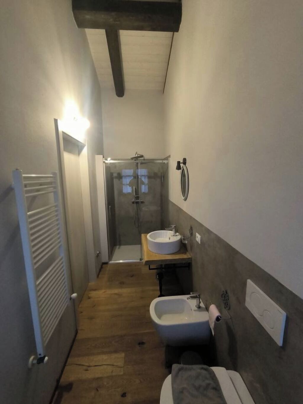 A narrow bathroom with a wooden floor, a glass shower enclosure, a round sink, and a bidet.