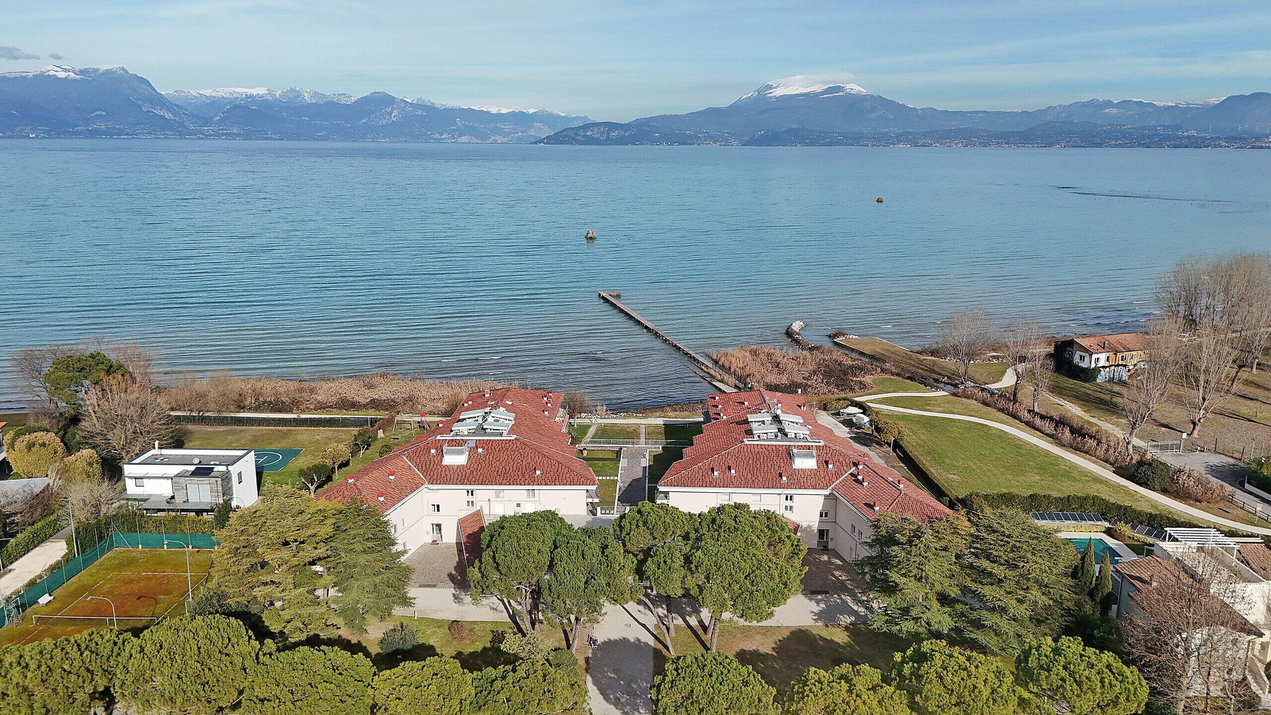 A residential area with houses having red-tiled roofs, a dock extending into a lake, and mountains in the background.