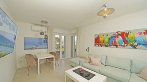 A modern living room with a light green sofa, a white coffee table, and a dining area with a white table and two chairs. A colorful parrot painting adorns the wall, and a large window provides a view of the outdoors. - Sunset on Sirmione in Sirmione (Sirmione)