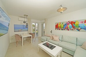 Ein modernes Wohnzimmer mit einem hellgrünen Sofa, einem weißen Couchtisch und einem Essbereich mit Tisch und zwei Stühlen. Ein farbenfrohes Papageienbild ziert die Wand, und ein großes Fenster bietet Blick ins Freie.