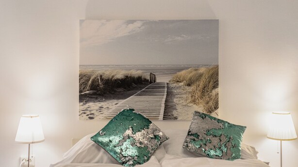 Ein Schlafzimmer mit großem Wandbild zum Strandthema, zwei Nachttischlampen und einem Bett mit weißer Tagesdecke.