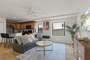 Une cuisine moderne avec des meubles en bois, un coin repas avec deux chaises, un canapé gris agrémenté de coussins à motifs, une table basse ronde et un ventilateur de plafond.