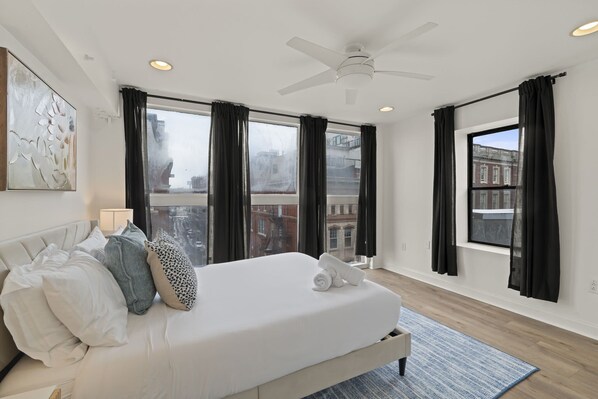 Ein Hotelzimmer mit Bett, Deckenventilator, großen Fenstern mit Vorhängen und Blick auf die Stadt.