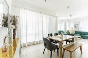 A modern living room with a dining area, featuring a large window with sheer curtains, a green sofa, and a wooden dining table with chairs. - HAVN - 1 bedroom in Dubai Marina with Marina Views (Dubai)