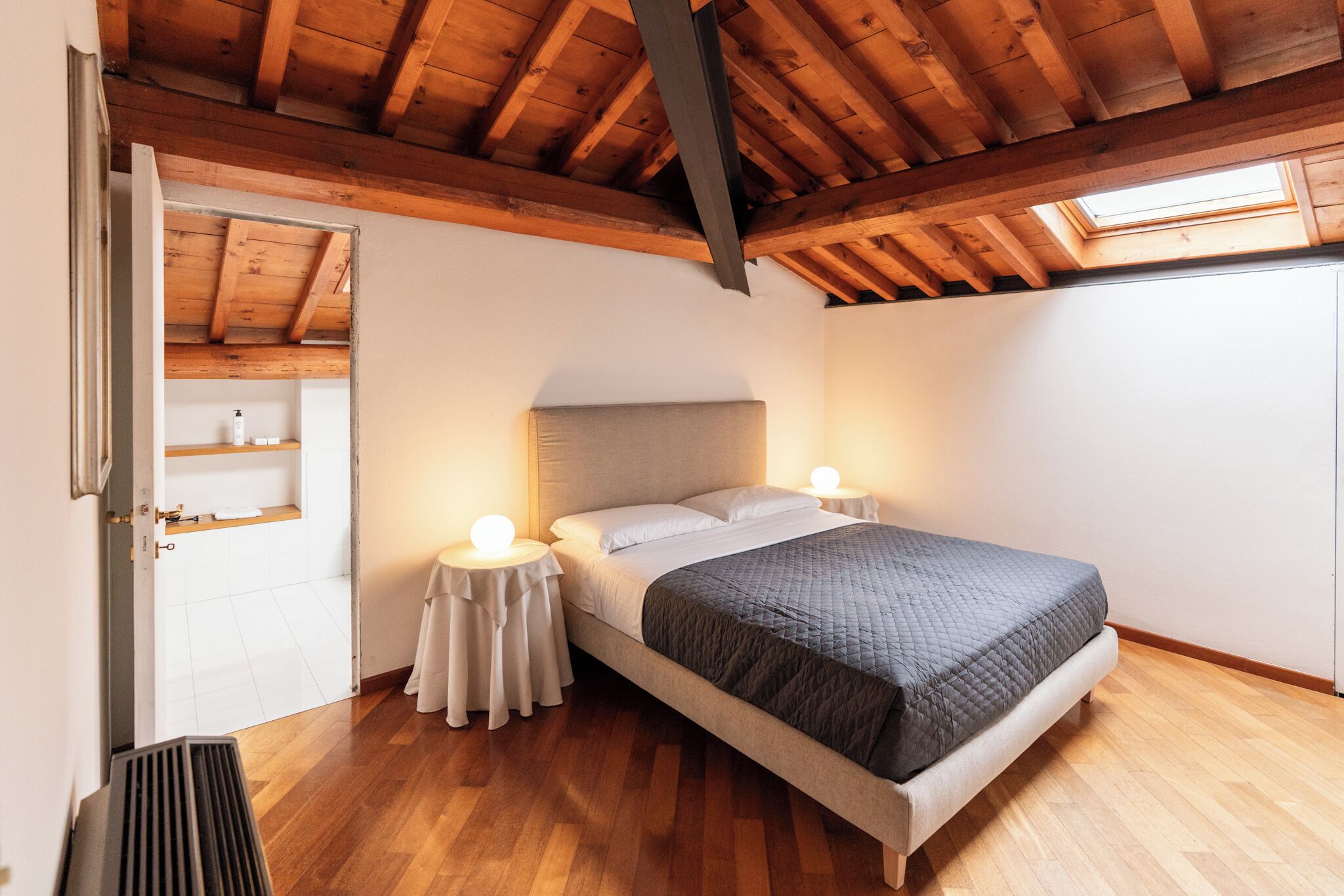 Ein Schlafzimmer mit Holzbalkendecke, einem Oberlicht, einem Bett mit grauer Bettdecke, Nachttischen mit Lampen und einer weißen Tür, die ins Badezimmer führt.