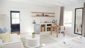 Een moderne, lichte woonkamer met een bank, salontafel en een eethoek met een tafel en stoelen. Er is een keuken met een spoelbak en een koelkast.