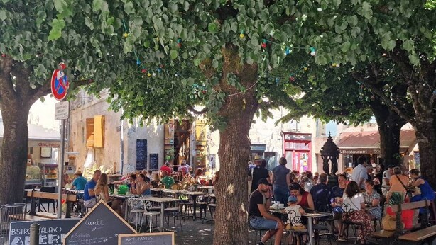 An outdoor café with tables and chairs under trees, surrounded by buildings and signs.