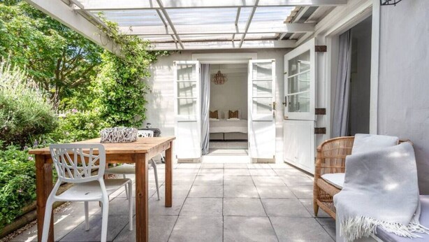 Eine sonnendurchflutete Terrasse mit einem Holztisch und -stühlen, einem Korbsessel und einer Glastür zu einem Innenbereich.