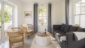 Ein modernes Wohnzimmer mit einem grauen Sofa, Korbstühlen, einem runden Couchtisch und großen Fenstern mit Vorhängen.