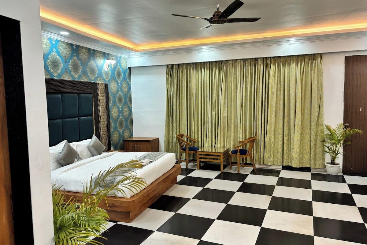 Quarto de hotel com piso xadrez, cama com cabeceira de madeira, ventilador de teto e uma janela grande com cortinas verdes.