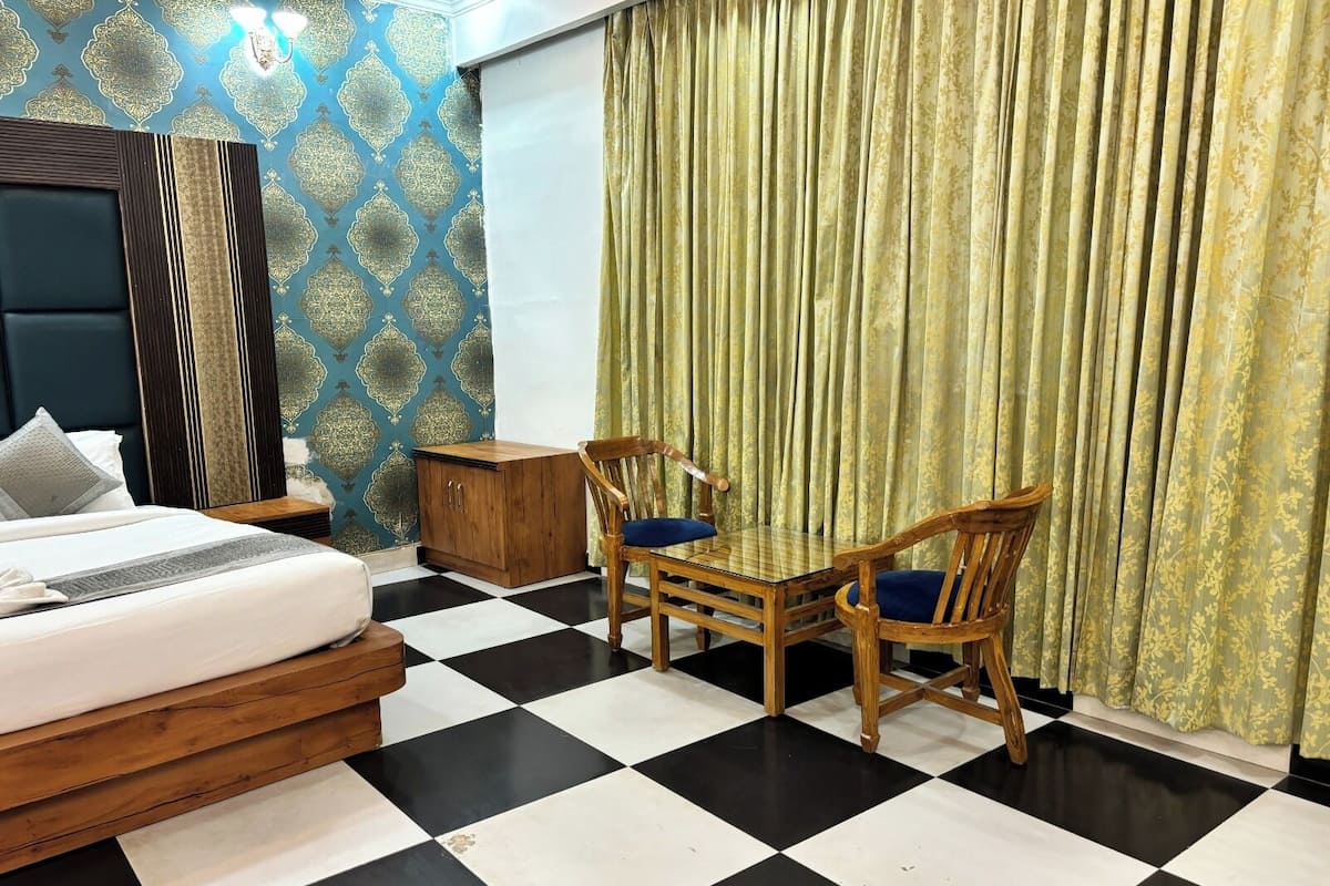 Quarto de hotel com piso xadrez, cama com cabeceira de madeira, armário de madeira, mesa com duas cadeiras e cortina com padrão floral.