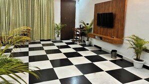 Una habitación con piso a cuadros blanco y negro, un mueble bajo de televisión de madera, un televisor de pantalla plana, una puerta de madera oscura y dos plantas en macetas.