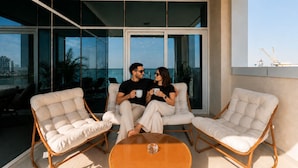 Un balcon moderne avec deux fauteuils confortables, une table ronde en bois et de grandes fenêtres du sol au plafond offrant une vue sur la ville.