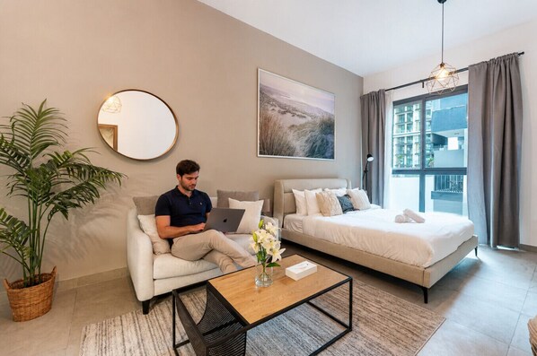 Ein modernes Hotelzimmer mit einer Couch, einem Bett, einem runden Spiegel, einem Gemälde und Blick auf Gebäude durch das Fenster.