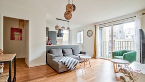 En moderne stue med en grå sofa, et lille rundt bord og en grøn lænestol. Der er et træspisebord med stole, et køkkenområde med en rød hvidevare og et stort vindue med udsigt til bygninger.