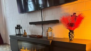 A modern living room with a fireplace, a flat-screen TV mounted on a white brick wall, and a decorative vase with red feathers. - Red Silence I Design Suite near Koblenz I Experience, Not Standard (Bendorf)