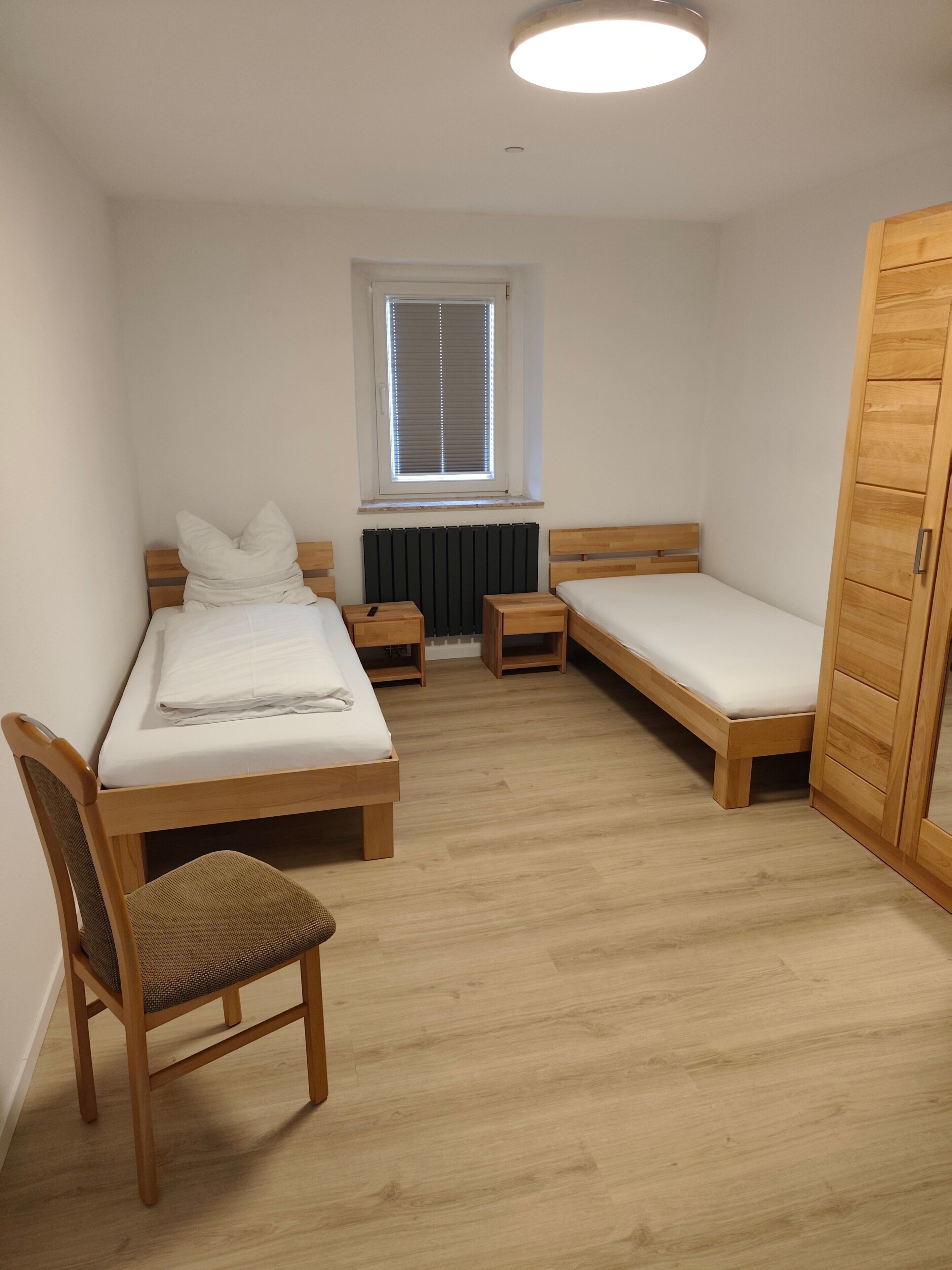 A small, tidy room with a single bed, a chair, a bedside table, a window with blinds, and a wardrobe.