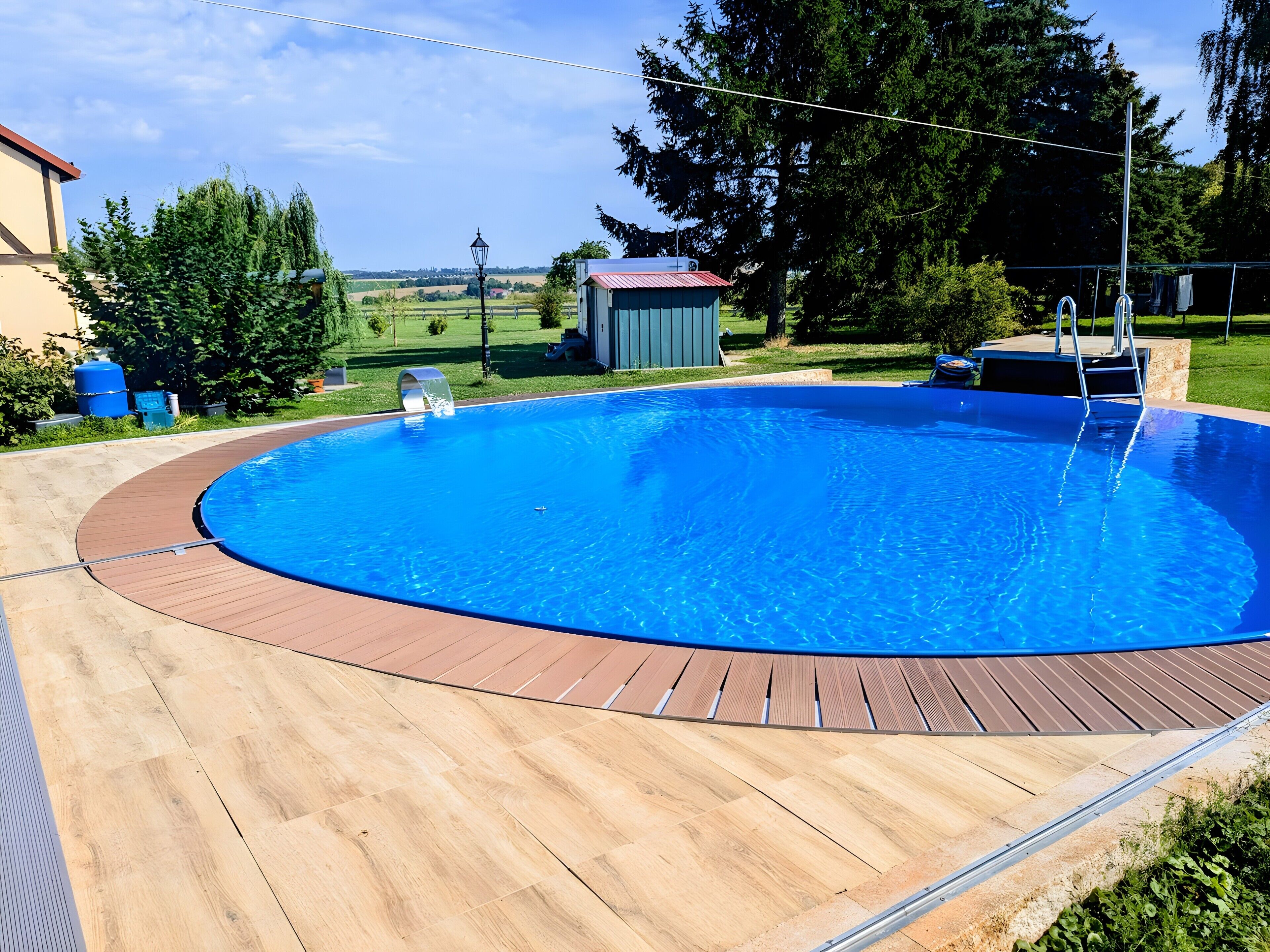 A circular swimming pool with a wooden deck, surrounded by greenery and a clear sky.