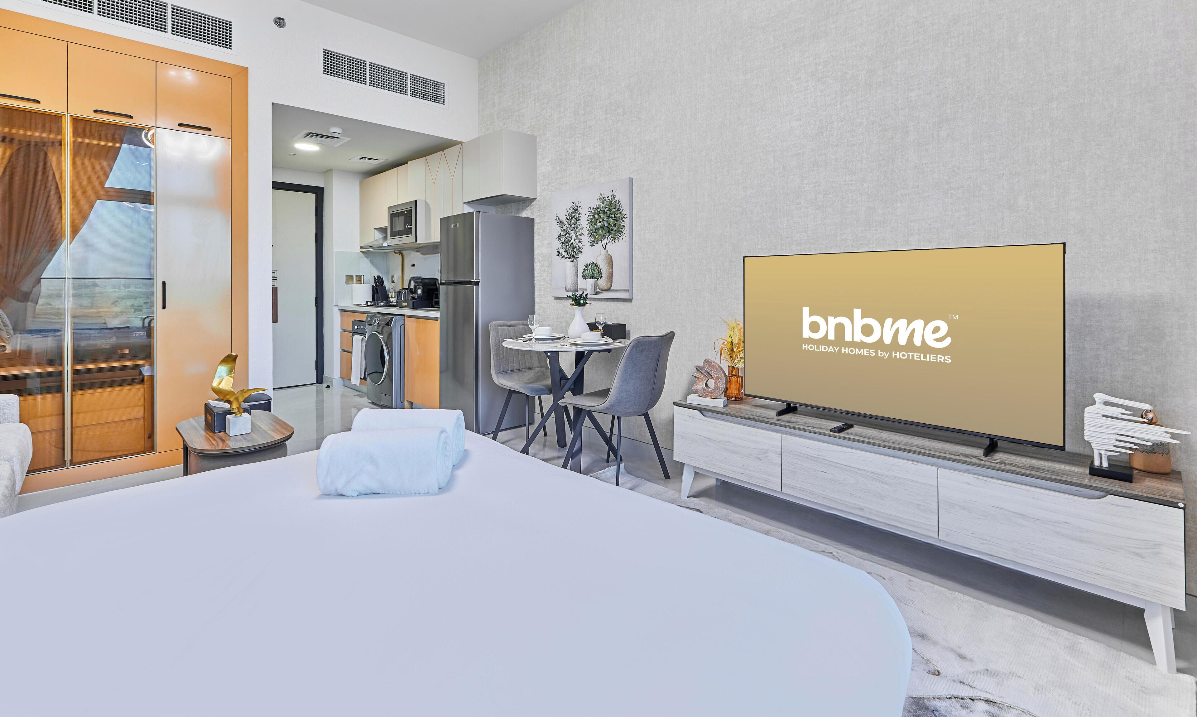 Habitació d'hotel moderna amb un llit gran, zona de menjador amb dues cadires, cuina i televisió que mostra 'bnbme'.