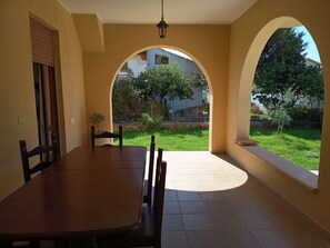 A dining area with a table and chairs, an arched window, and a view of a garden and buildings. - Sardinian Wind House, vacation home South Sardinia 600 m from the sea (La Maddalena)