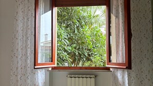Ein Zimmer mit Bett, einem Fenster mit geöffneten Lamellen und Heizkörper, mit Blick auf Grünfläche.