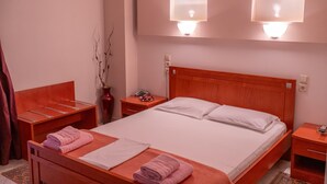 Une chambre d’hôtel avec un lit en bois, deux tables de chevet, une petite table d’appoint, une lampe fixée au mur et un vase contenant des branches séchées.