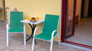 Un petit espace extérieur aménagé avec deux chaises vertes et une table blanche, où l’on aperçoit une assiette de croissants et deux verres de jus d’orange.