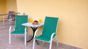 A small outdoor seating area with two green chairs and a table, a plate of fruit, and two glasses of orange juice.